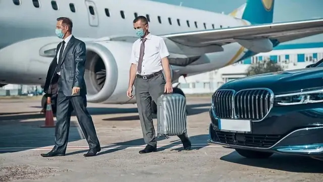 A driver waiting beside a car on the airport apron as passengers arrive with luggage